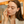 A person with long brown hair gently applying white whipped tallow balm to their cheek as part of their skincare routine. They are wearing a light beige or cream-colored robe over a dark top, and their eyes are closed in a peaceful expression. The balm appears creamy and smooth on their skin. The warm-toned bathroom setting features wooden cabinetry and soft lighting, creating a serene self-care atmosphere.