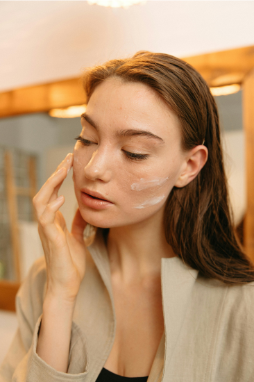 A person with long brown hair gently applying white whipped tallow balm to their cheek as part of their skincare routine. They are wearing a light beige or cream-colored robe over a dark top, and their eyes are closed in a peaceful expression. The balm appears creamy and smooth on their skin. The warm-toned bathroom setting features wooden cabinetry and soft lighting, creating a serene self-care atmosphere.